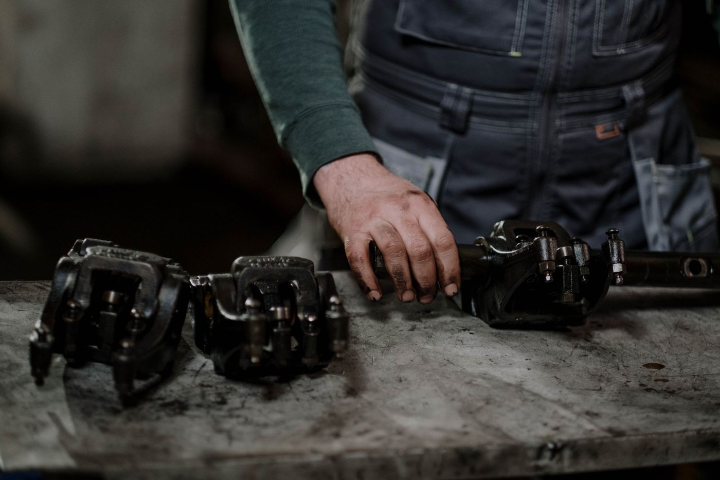 Close-up of a mechanic's greasy hand resting on a used suspension rail, with two salvaged brake calipers laid out beside it on a grimy steel bench