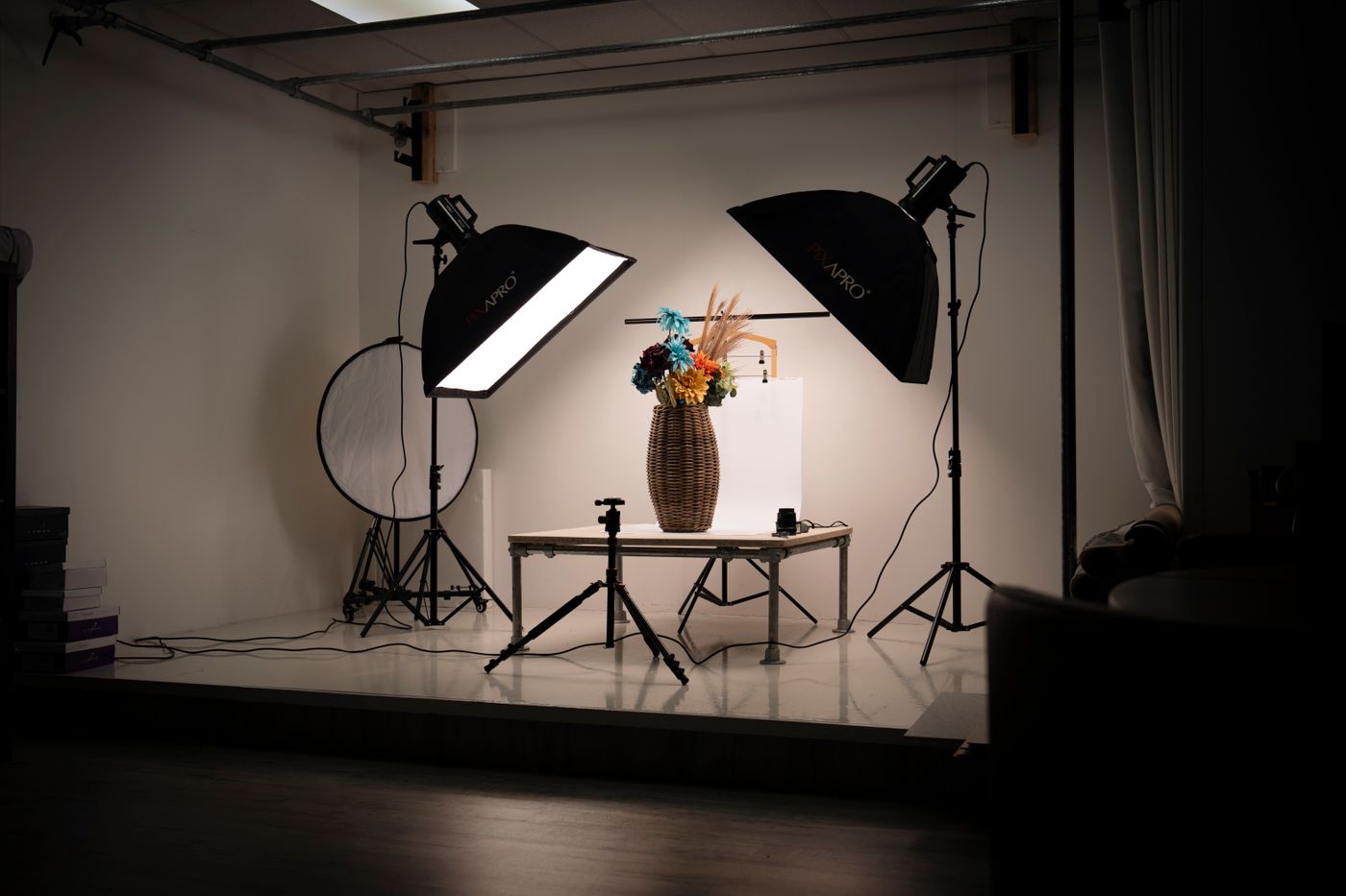 An indoor photography studio setup: a softbox on a light stand and a tripod arranged around a floral arrangement standing in a basket