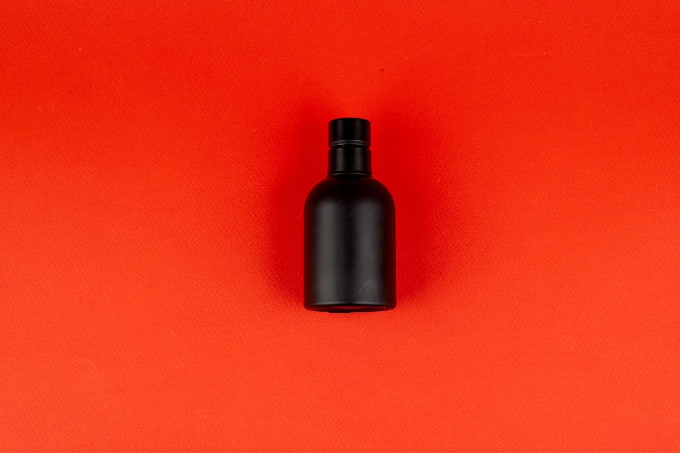 A matte black cylindrical bottle photographed from above, centred on a flat, evenly lit bright red backdrop.