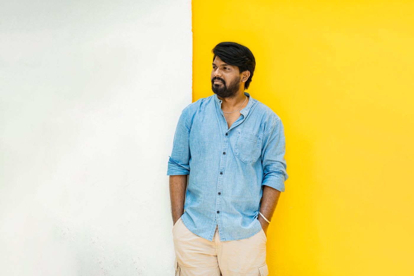 Bearded man in a denim shirt standing against a wall that is flat yellow on one side and plain white on the other, with empty space beside him