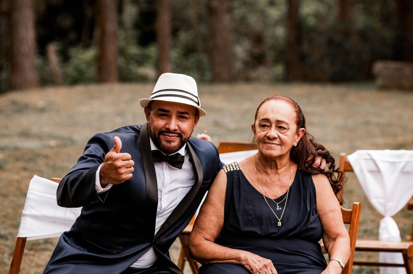 An elderly woman in a black dress sits at an outdoor wedding while a man in a tuxedo and hat leans in with his arm behind her shoulder.