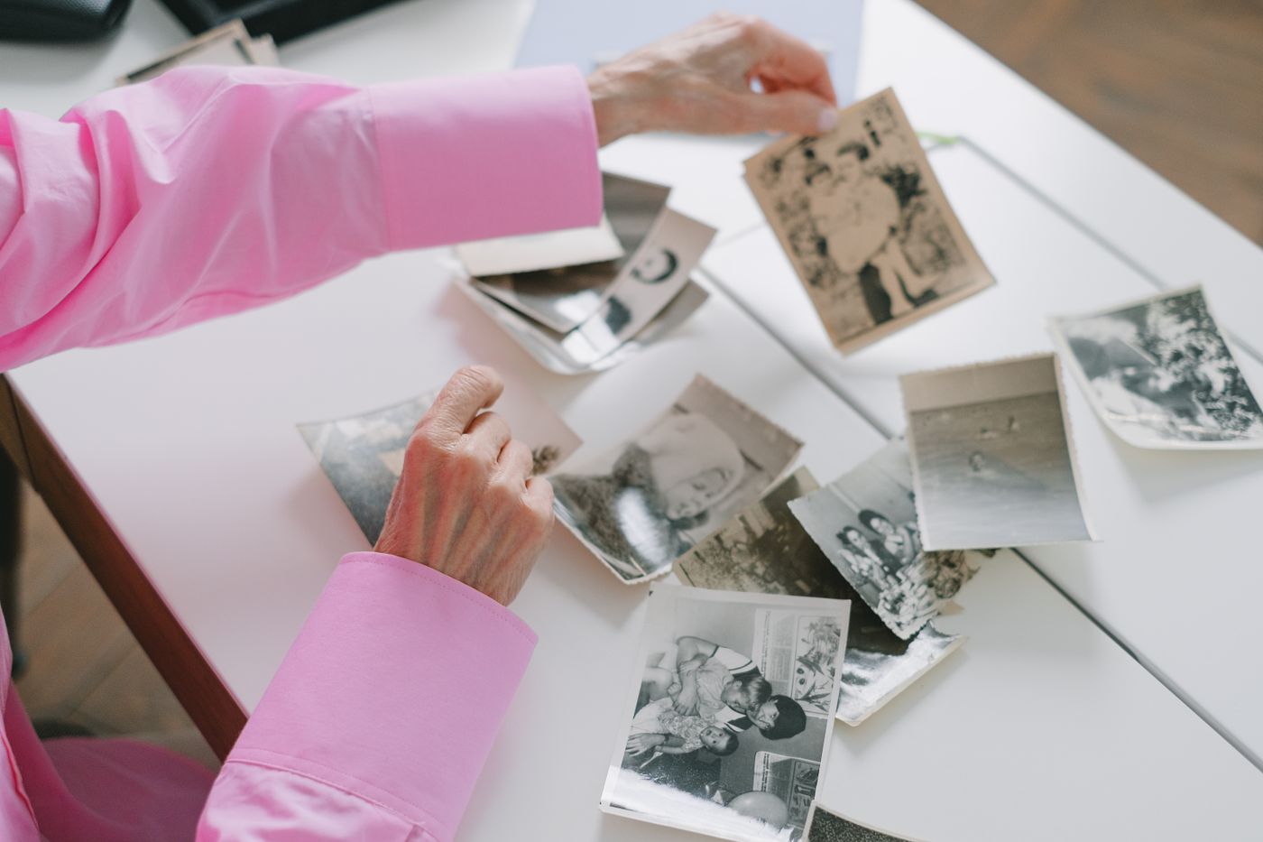 An older woman in a pink blouse sorts through dozens of loose black-and-white family photographs spread across a white table.