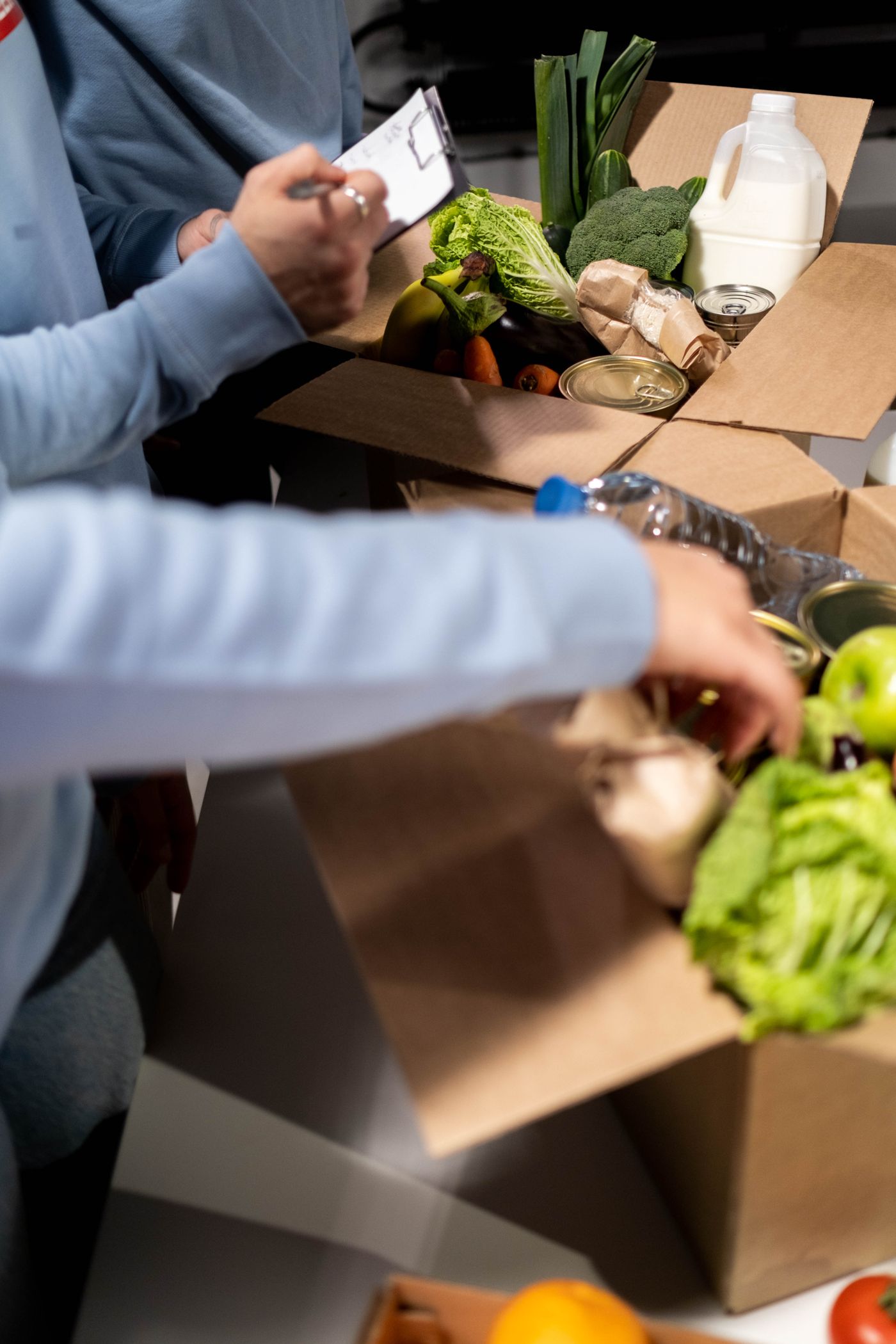 Two volunteers in blue sweatshirts filling cardboard boxes with vegetables, bread, tinned food and milk, framed so only their hands and sleeves are visible