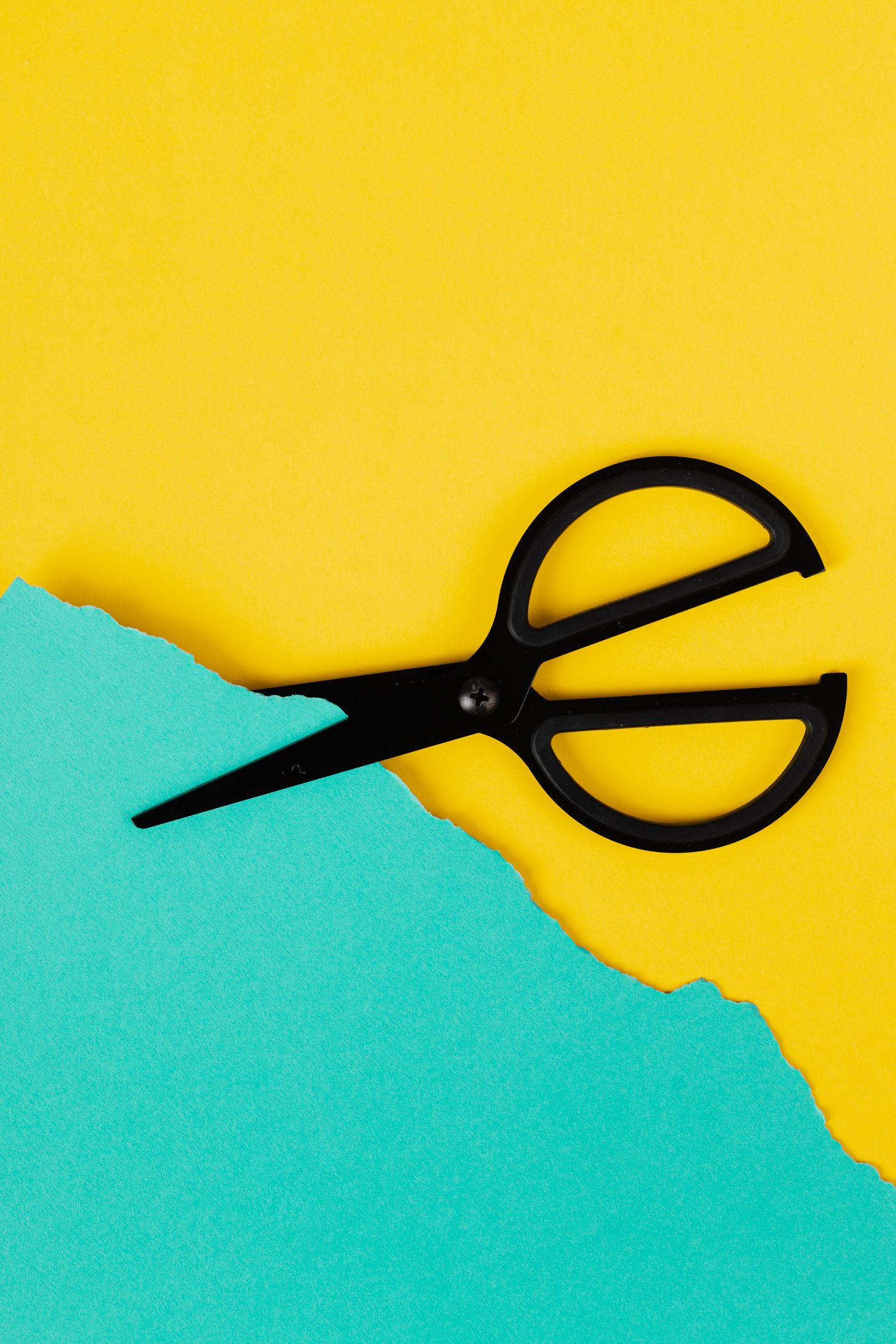 A pair of black-handled scissors resting closed on a bright yellow sheet beside torn turquoise paper, the yellow showing plainly through both finger loops