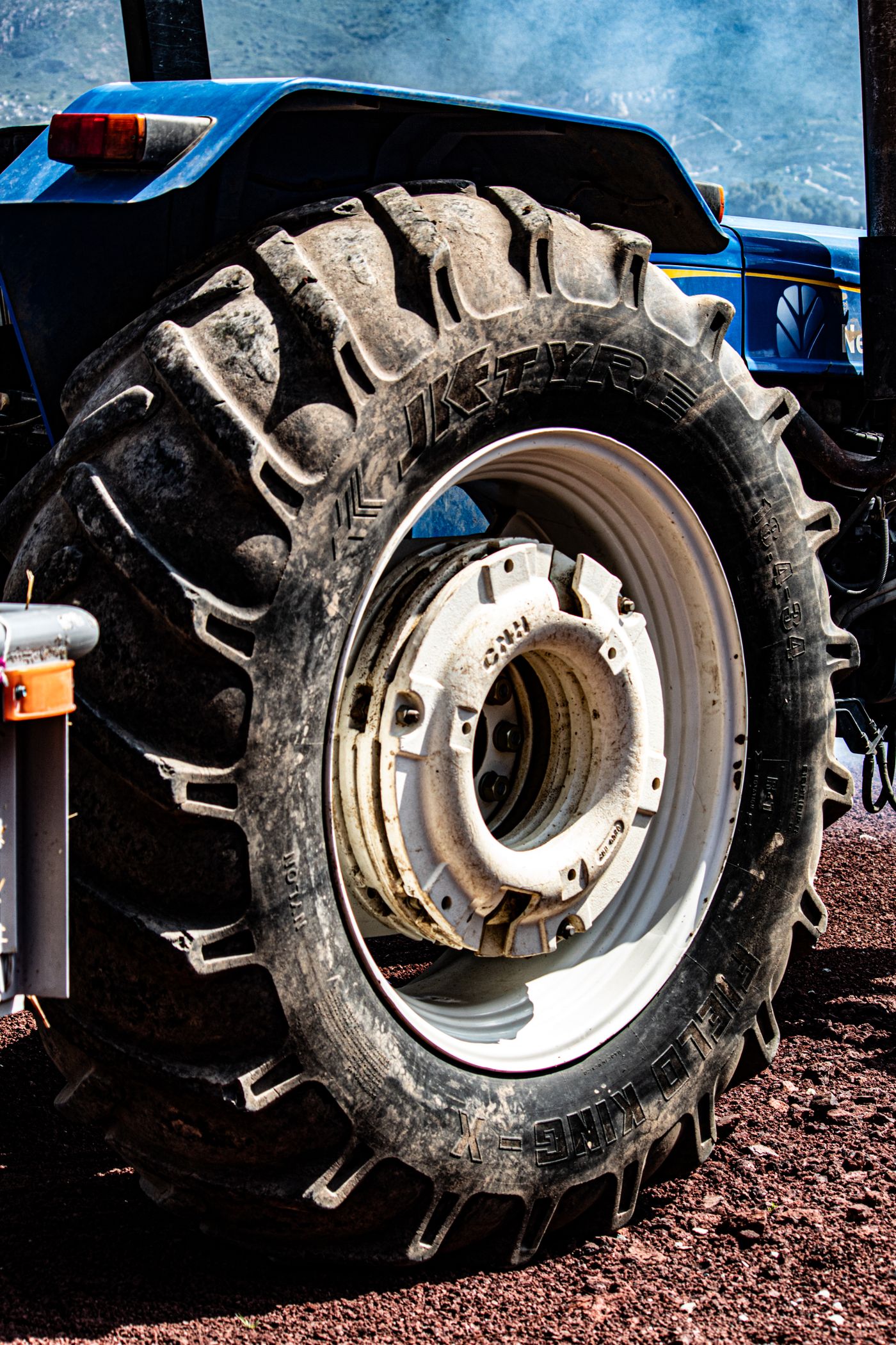 Nahaufnahme eines staubigen Traktor-Hinterreifens von der Seite: Stollenprofil, Flankenbeschriftung, zerkratzte weiße Felge