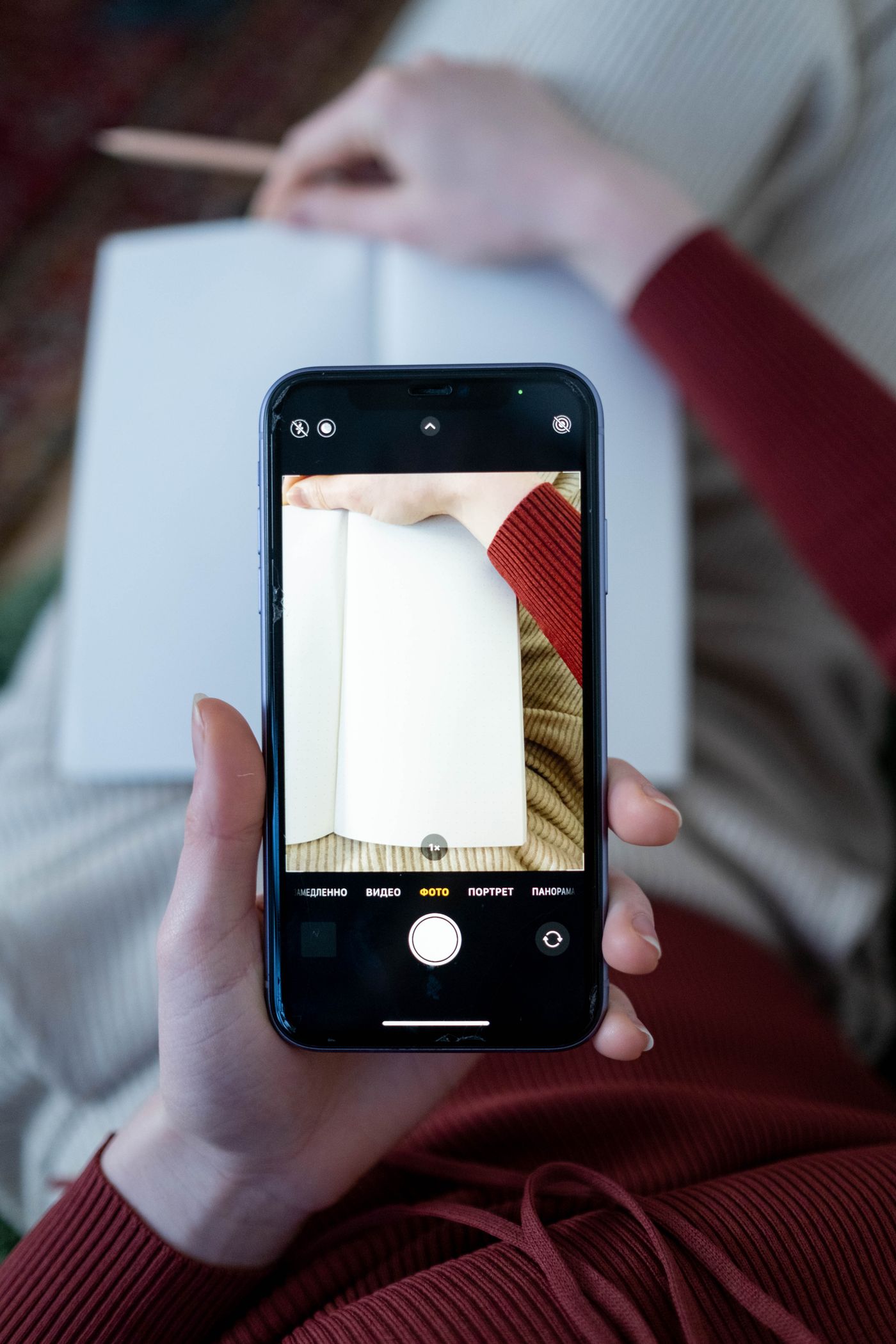 A person holding a smartphone with the camera app open, framing a blank white notebook page lying flat below it