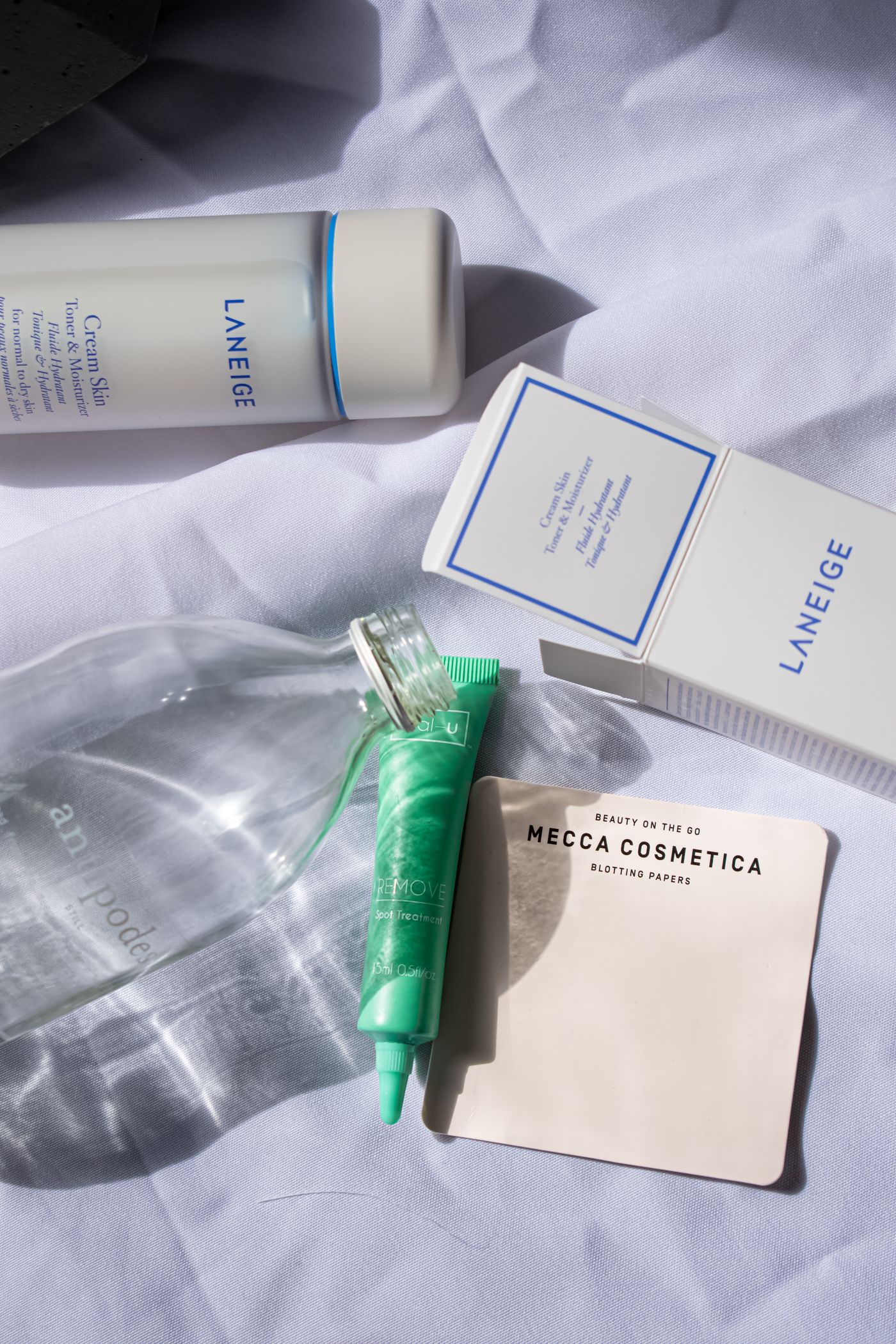 Flat lay на козметичен комплект върху светлосин плат, където зелена туба лежи върху прозрачна стъклена бутилка, зад тях стои картонена кутия с печат, а под тубата е подпъхнато саше