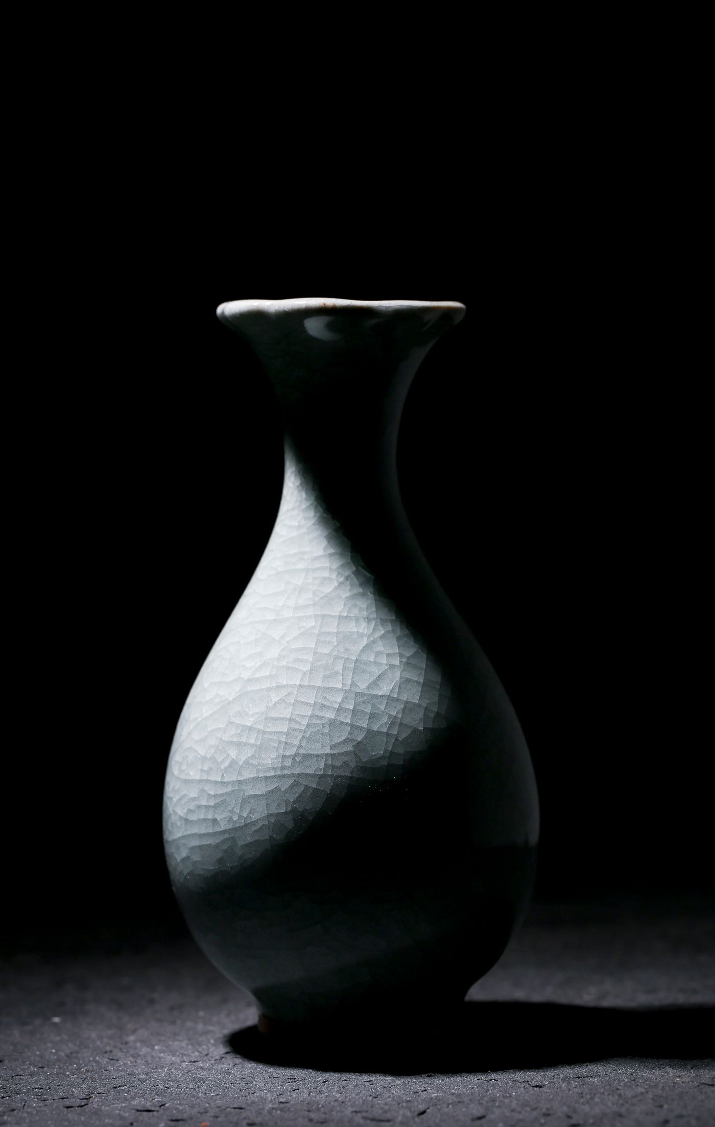 A pale crackle-glazed ceramic vase lit against a near-black background, its silhouette separated from the backdrop purely by brightness with almost no colour present.
