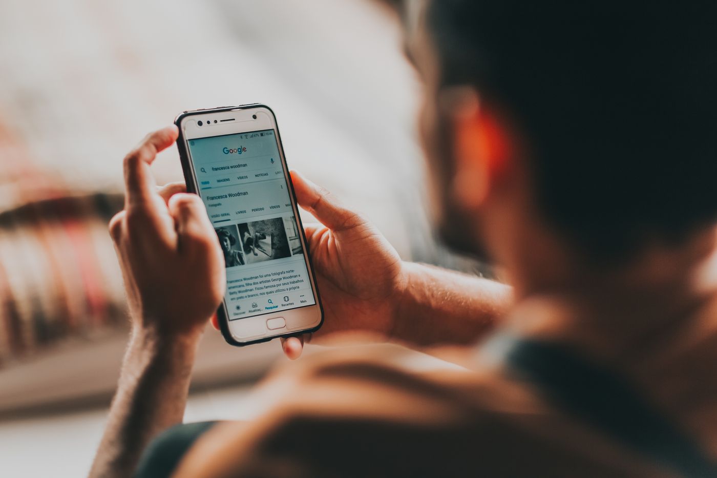 Über-die-Schulter-Blick auf einen Mann, der ein Smartphone in beiden Händen hält und auf dem Display eine Webseite mit kleinen Vorschaubildern liest