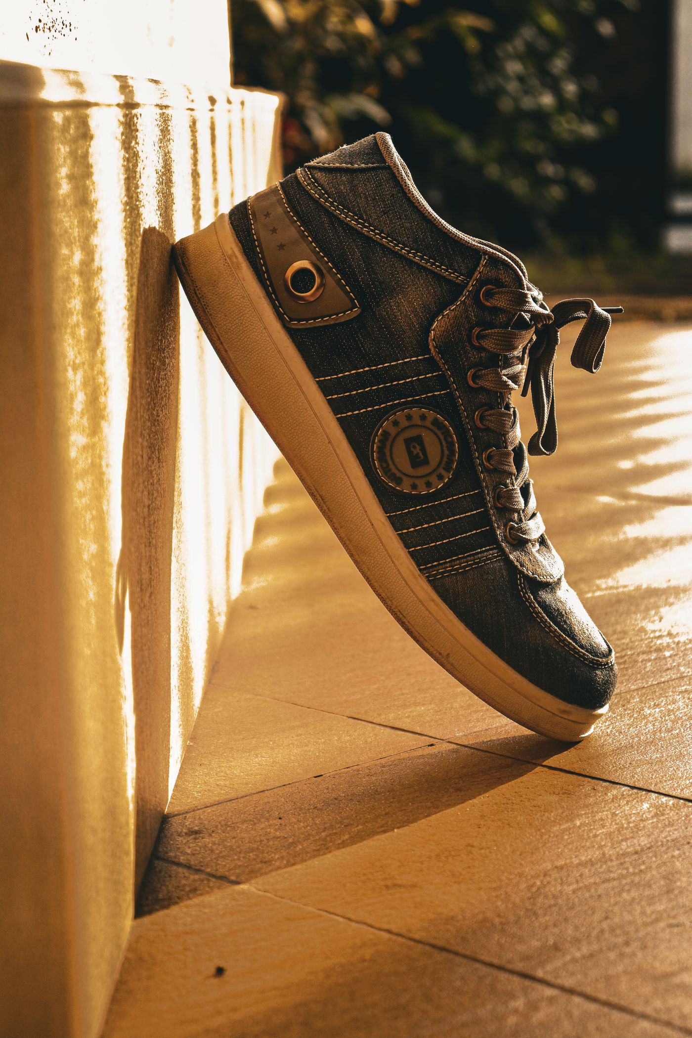 A denim sneaker propped upright with its heel touching a pale wall in warm low sunlight, its shadow pooling right where the shoe meets the surface