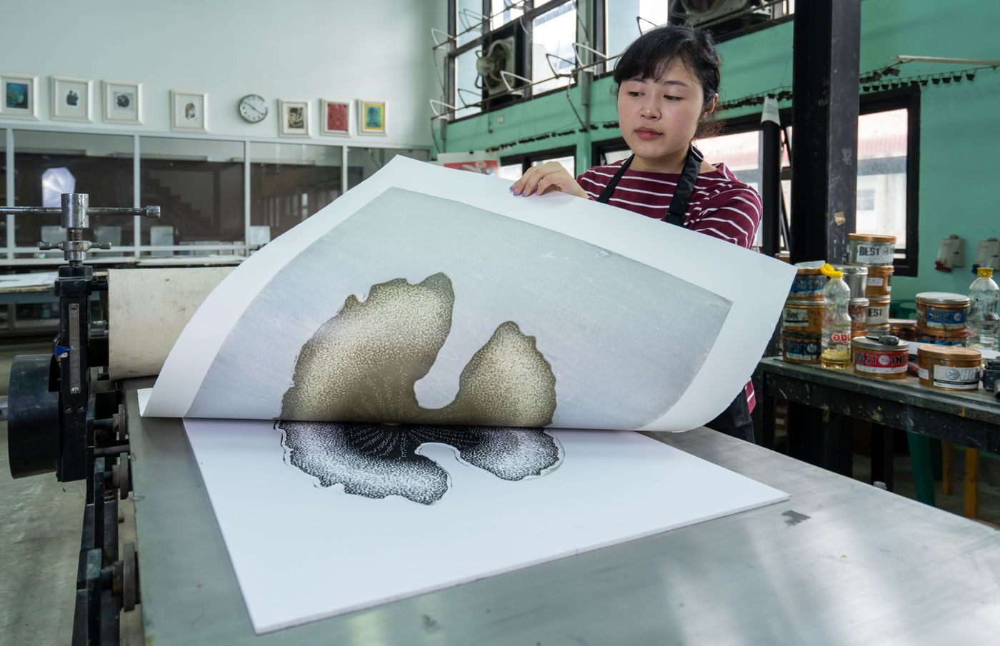 A woman in a print studio holding a freshly printed sheet up to the light and examining the edges of the artwork closely.