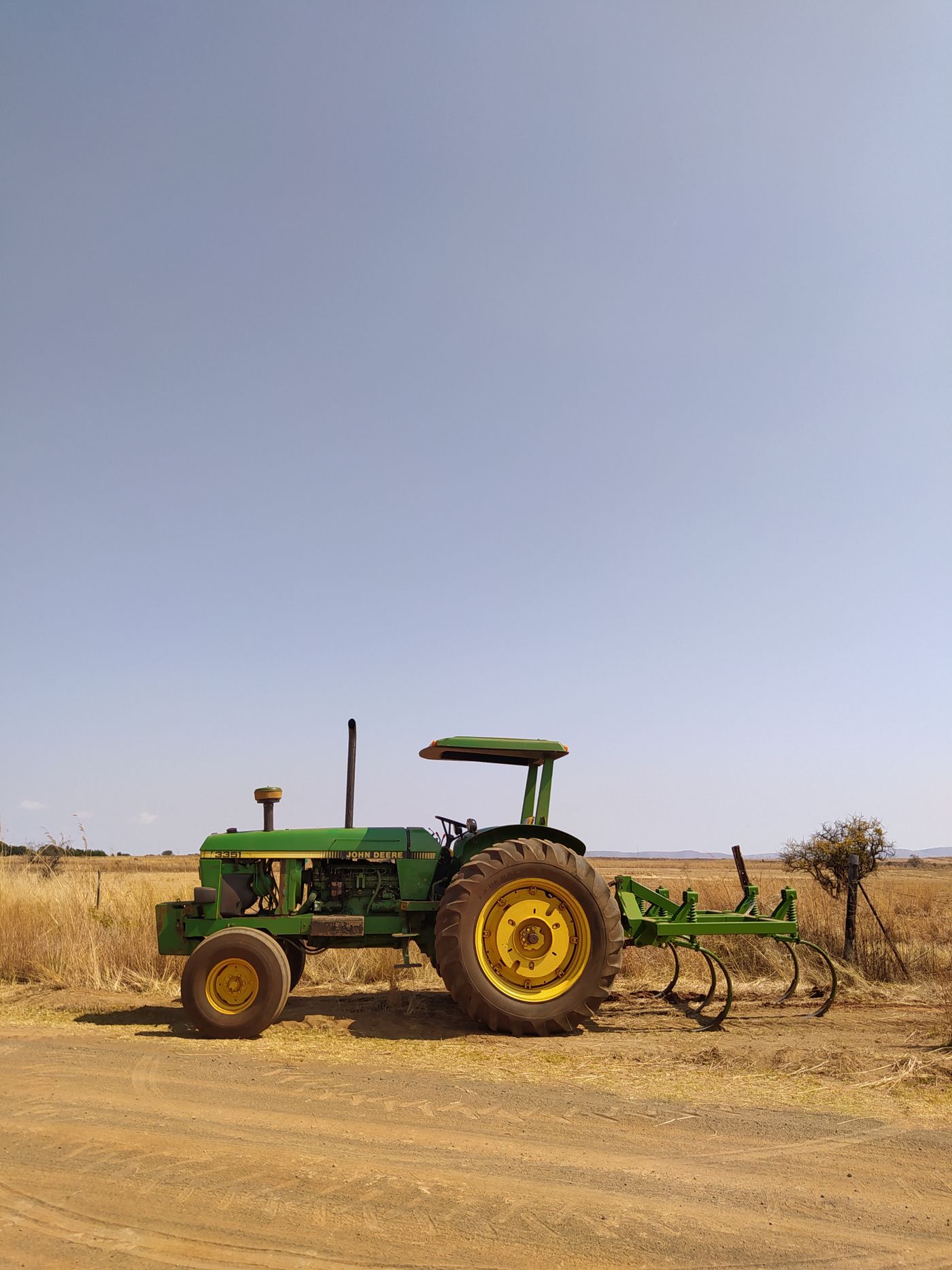 Grüner Traktor mit angebautem Grubber, seitlich am Feldweg geparkt, dahinter offener Himmel und trockenes Grasland in harter Mittagssonne