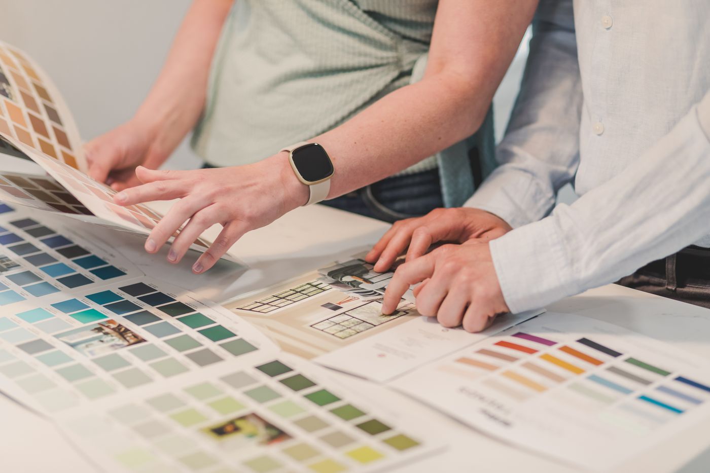 Two people leaning over a desk covered with printed colour sample charts and a swatch booklet, one of them pointing at a chart while the other turns a page.